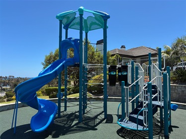 Blue playground structure with a spiral slide, climbing platforms, and stairs in an outdoor park on a sunny day.