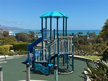 Blue and gray playground climbing structure with slides and railings, overlooking the ocean on a clear sunny day.