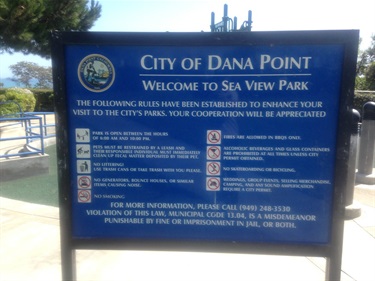 Blue sign reading “City of Dana Point – Welcome to Sea View Park” with park rules listed below, mounted on black posts in front of a playground area.