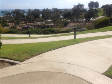 Curving concrete pathways in a grassy park overlooking trees, landscaping, and the ocean in the distance under a partly cloudy sky.