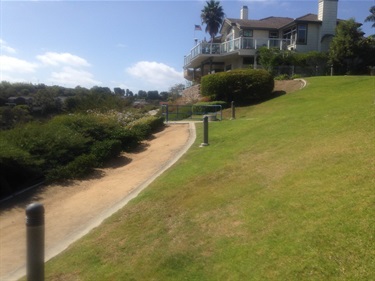 Dirt pathway bordered by grass and landscaping, leading uphill toward a large house with a balcony and palm trees under a clear blue sky.