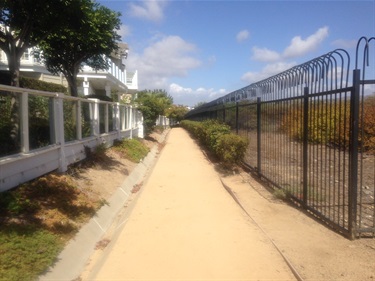 Dirt pathway bordered by a white fence on the left and a black metal fence on the right, with shrubs and trees along both sides under a partly cloudy sky.