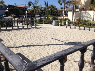 Square sand play area enclosed by a weathered wooden railing with rope accents, surrounded by palm trees, shrubs, and nearby houses under a clear blue sky.