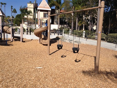 Two swings on a wooden swing set in a playground with castle-themed play structures and slides on a wood chip surface, surrounded by palm trees and houses under a clear blue sky.