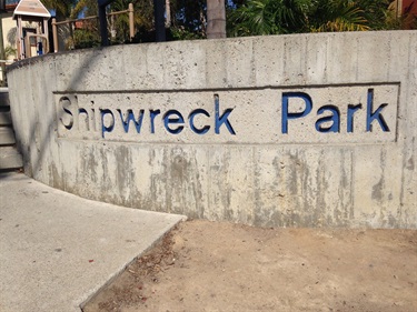 Sand play area bordered by a wooden railing with rope accents, with castle-themed playground structures in the background and a street intersection visible beyond under a clear blue sky.