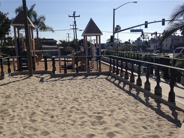 Sand play area bordered by a wooden railing with rope accents, with castle-themed playground structures in the background and a street intersection visible beyond under a clear blue sky.
