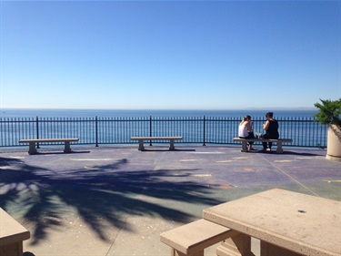 Two people sitting on benches facing the ocean, on a paved lookout area with a metal railing, under a clear blue sky with calm water in the background.