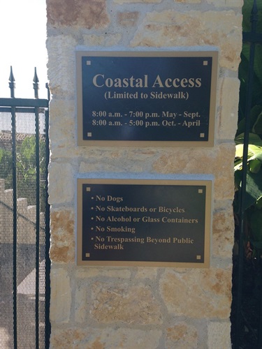 Stone pillar with two metal signs reading “Coastal Access” and listing hours and rules. Rules include no dogs, skateboards, bicycles, alcohol, smoking, or trespassing beyond sidewalks.
