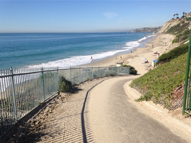 Curved paved path descending toward Strands Beach with a metal fence on the left. Ocean waves and coastline stretch into the distance under a clear blue sky.