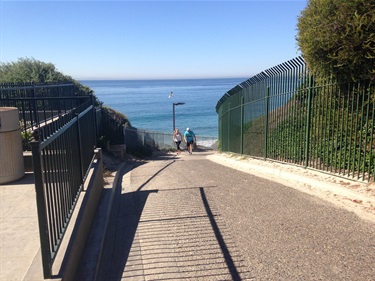 Steep paved path leading down toward the ocean, bordered by tall green metal fences on both sides. Two people are visible near the bottom, with clear blue water and sky in the background.