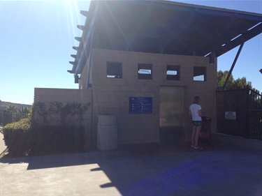 Concrete restroom building with blue doors and angled roof, surrounded by trash bins and railings, overlooking the ocean under a clear blue sky.