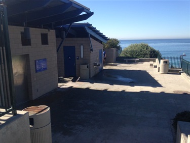 Concrete restroom building with blue doors and angled roof, surrounded by trash bins and railings, overlooking the ocean under a clear blue sky.