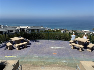 Strands Vista Park coastal overlook with picnic tables and benches on a painted concrete surface, facing a fence-lined edge with ocean views and rooftops below under a clear blue sky.