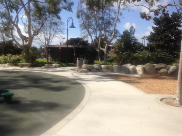 Curved concrete pathway in a park with trees, large rocks, and landscaped areas, including a green play surface and a lamppost in the background.