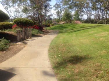 Curved concrete pathway through a landscaped park with green grass, shrubs, and trees under a partly cloudy sky.