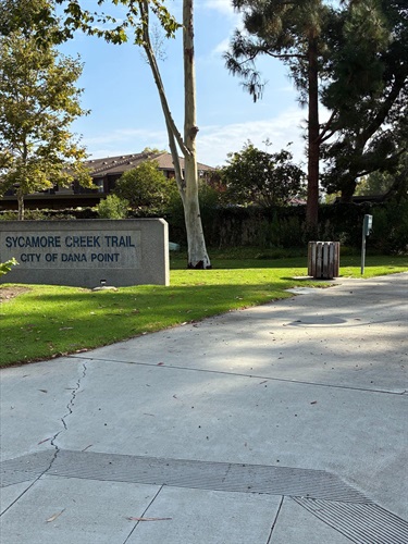 Entrance to Sycamore Creek Trail in Dana Point, with a stone sign, green lawn, trees, and a paved walkway.