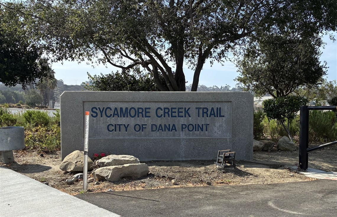 Sycamore Creek Trail sign at a trail entrance in Dana Point, surrounded by rocks, trees, and natural landscaping.