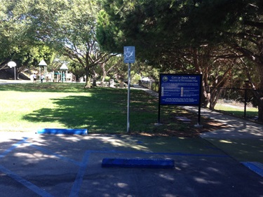 Thunderbird Park entrance area with a blue informational sign and a handicap parking space in the foreground, trees providing shade, and a playground visible in the background.