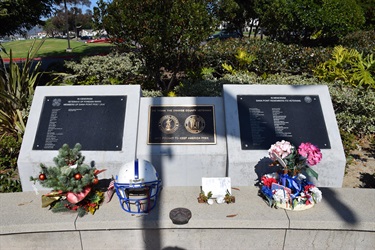 Memorial display with three plaques mounted on a stone base, featuring engraved names and military emblems, surrounded by flowers, a football helmet, and tribute items in an outdoor park setting.
