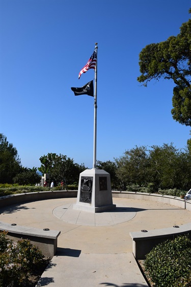 Outdoor memorial featuring a tall flagpole with the U.S. flag and POW/MIA flag, mounted on a stone base with plaques, surrounded by a circular paved area and landscaped greenery under a clear blue sky.