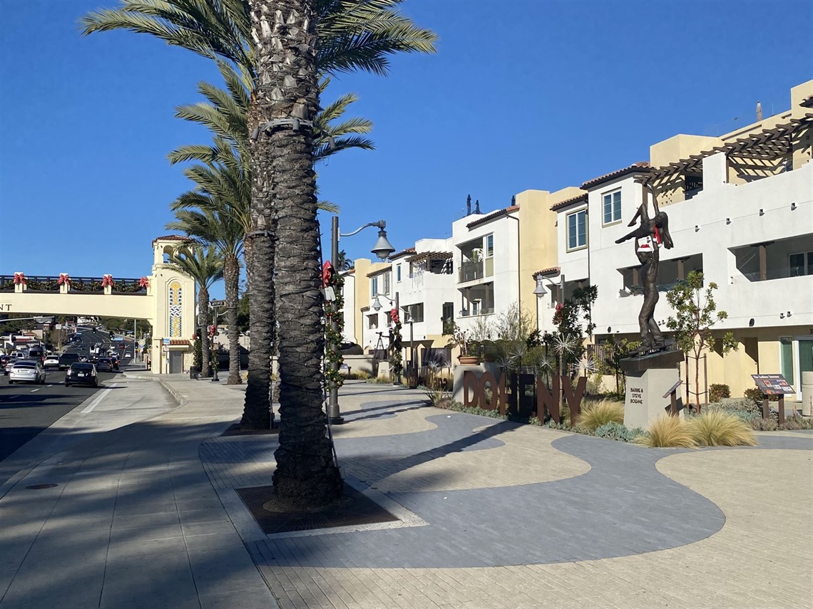 Watermen's Plaza promenade with tall palm trees lining a wide walkway, beside white multi-story buildings with balconies under a clear blue sky.
