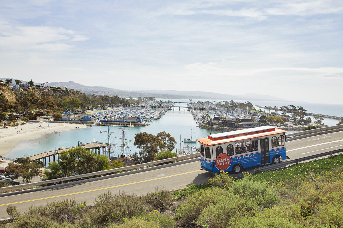 The Dana Point Trolley traveling along a coastal road with a marina full of boats and green hills in the background. The sky is partly cloudy.
