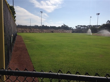Baseball field at Del Obispo Park with sprinklers watering the grass, viewed above a chain-link fence, with light poles and trees in the background under a partly cloudy sky.
