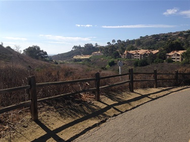 Scenic view of a paved path with a wooden fence, dry brush on the hillside, and resort buildings in the distance under a clear blue sky.