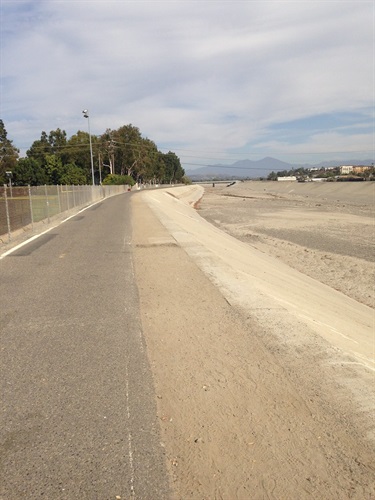 Wide paved path running alongside a sandy channel, with a fence and trees on the left and mountains visible in the distant background under a partly cloudy sky.