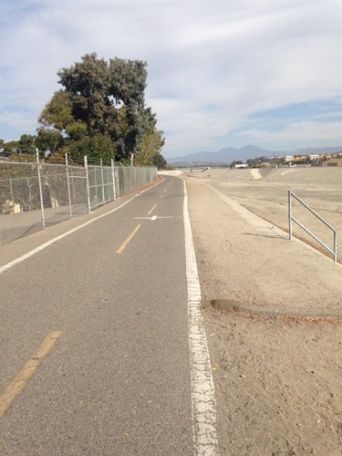 Paved bike path with a yellow center line next to a sandy channel and chain-link fence. Trees and distant mountains under a partly cloudy sky.