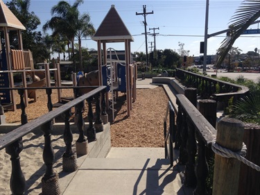 Playground area with wooden railings, ramps, and structures featuring peaked roofs, surrounded by mulch and palm trees near a street.