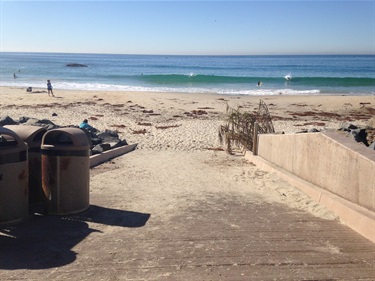Concrete ramp leading to Strands Beach with trash bins on the left. Ocean waves in the background with a few people near the shoreline under a clear blue sky.