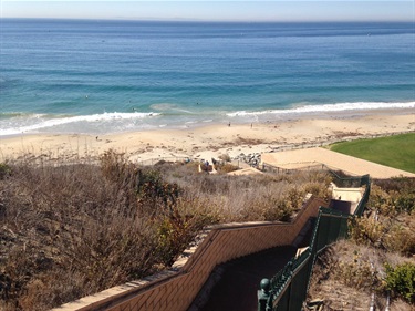View from a hillside showing a wooden stairway leading down to Strands Beach. Ocean waves in the background under a clear blue sky, with some greenery and a grassy area near the stairs.