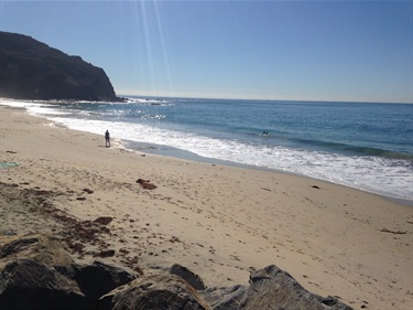 Strands Beach with gentle waves washing ashore, rocky cliff on the left, and a person walking near the water under a bright blue sky.