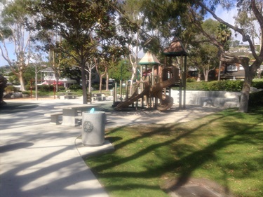 Playground with wooden climbing structure and slide surrounded by trees, concrete paths, benches, and grassy areas in a park setting.
