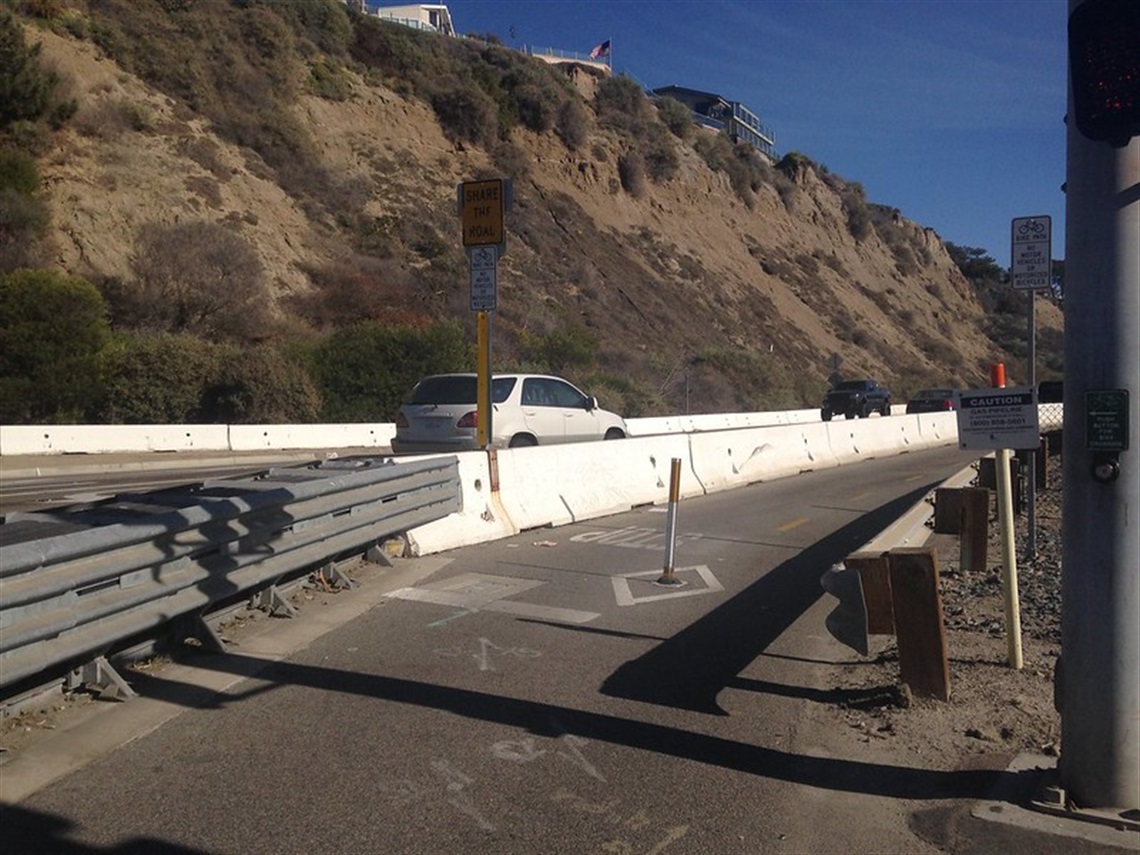 Coast Highway Trail with a white car driving on the left side of the barrier, and another vehicle can be seen in the distance. The road is situated next to a hillside with houses visible at the top. 