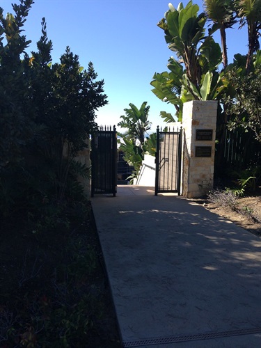 Open black metal gate between two stone pillars leading to a descending walkway. Lush green plants surround the entrance, with clear blue sky visible in the background.